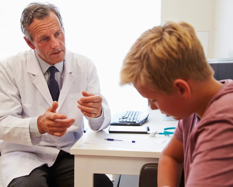 teenage boy listening to a male doctor wearing a white coat sat at a desk trying to get a CAMHS referral
