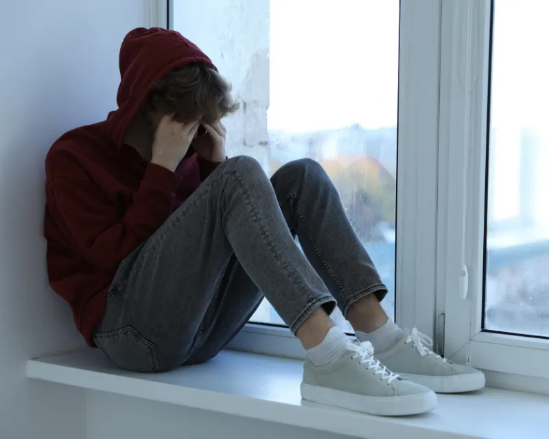A teenage boy sitting on a windowsill, his hoodie up and his hands over his face, showing signs of depression in teenagers