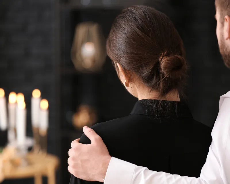 a man with his arm around the shoulders of a woman wearing black looking at lit candles representing grief and understanding how long it lasts and the timeline