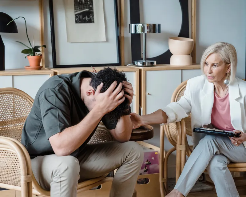 A man sat on a wicker chair with his head in his hands, with a woman therapist in a white blazer trying to console him, representing my teenager is making me depressed
