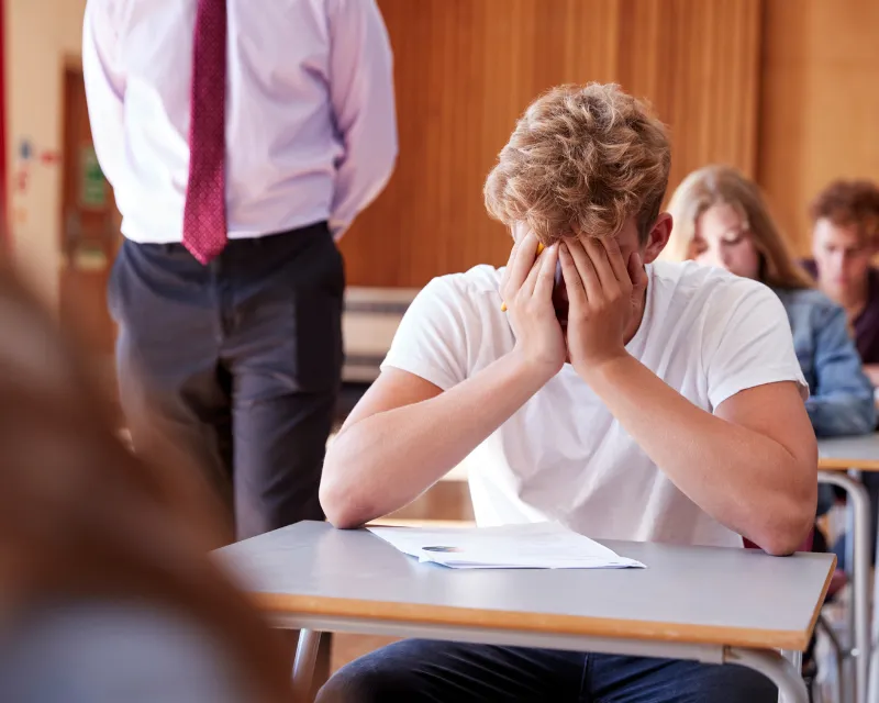 A teenage boy sitting an exam in a school hall with his hands over his face and a teacher walking up the aisles represents red flags in teenage behaviour