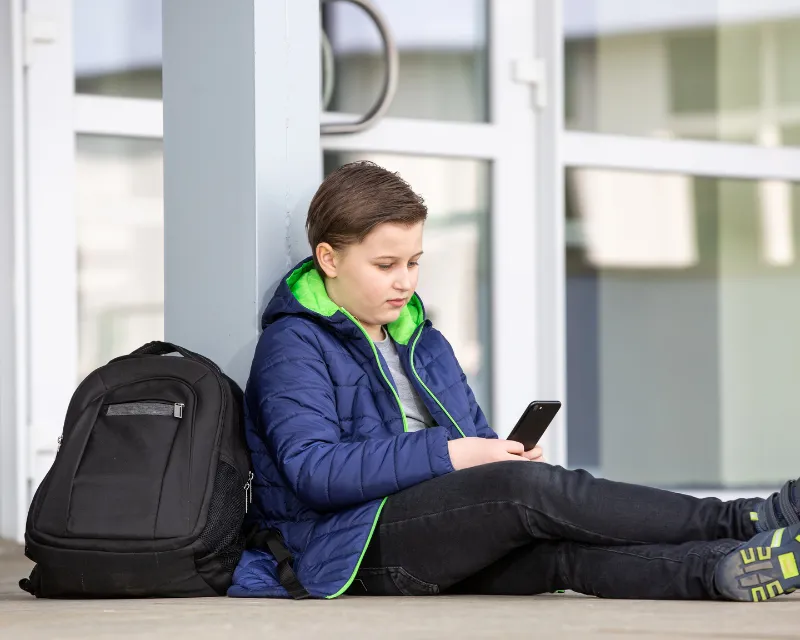 A young boy sat outside of a school with a coat on and a rucksack next to him, holding a phone, representing school refusal in young people