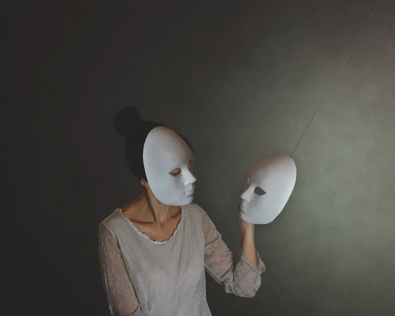 A woman stood in front of a dark background wearing a white mask and holding another white mask in her hand which she is looking at to represent ADHD masking