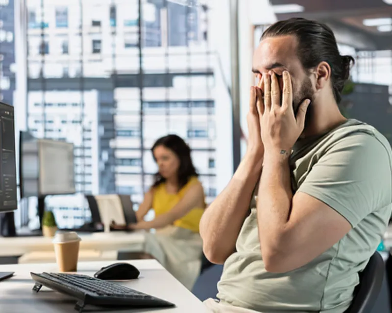 A man sat at a computer desk in a light office with a large glass window in teh background. He is sat with his hands covering his face showing signs of adult autistic burnout
