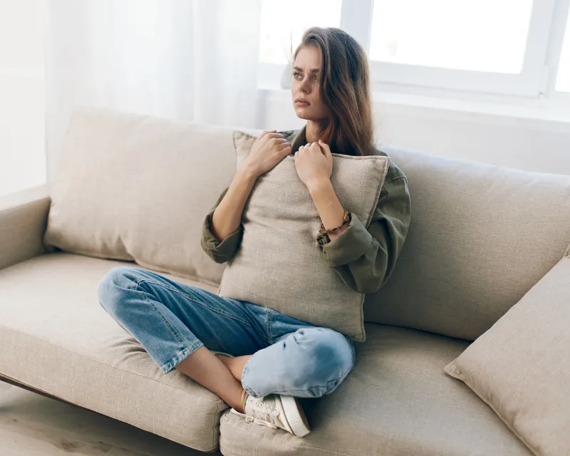 a young woman sat on a sofa hugging a pillow with her legs crossed and looking afraid and showing signs of grief and anxiety