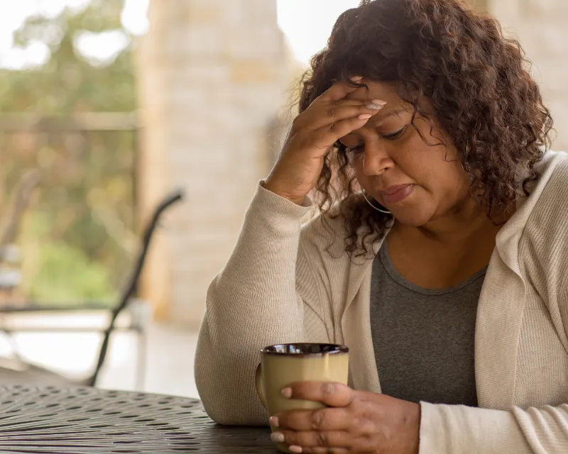 A woman sat at a garden table with a cup of coffee, staring into the cup with her hand on her forehead, showing signs of perimenopause anxiety